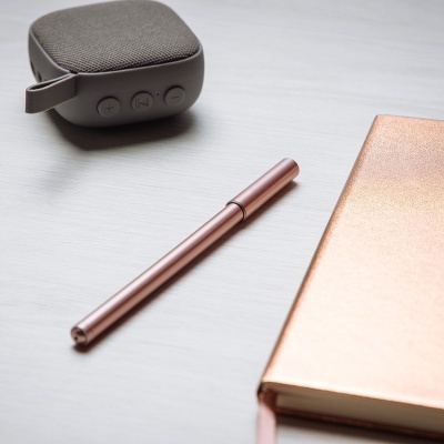 Coluna portátil cinza, caneta e agenda rose gold em superfície branca.