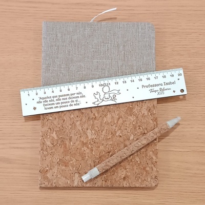 Caderno com capa de cortiça e tecido, régua branca com frase e nome, caneta de cortiça sobre mesa de madeira