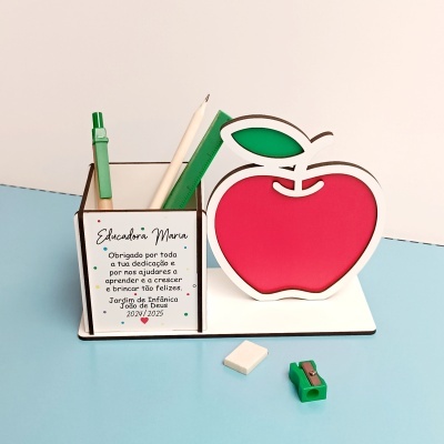 Porta-canetas branco personalizado com maçã de madeira vermelha, régua verde, caneta verde, lápis branco, apontador e borracha sobre mesa azul