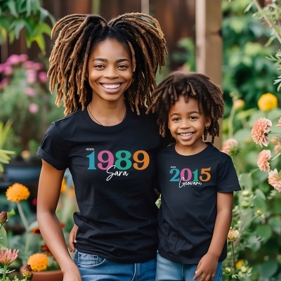 Mulher e criança com t-shirts pretas estampadas com anos e nomes, em jardim com flores coloridas.