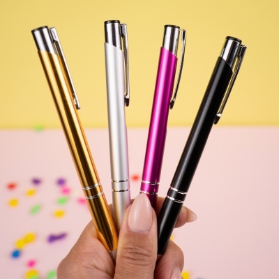 Quatro canetas de metal coloridas, dourado, prateado, rosa e preto, com clips em fundo amarelo e rosa