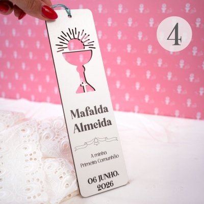 Marcador de livro branco com recorte de cálice e sol e texto impresso a preto, segurado por mão com unha vermelha sobre fundo rosa.