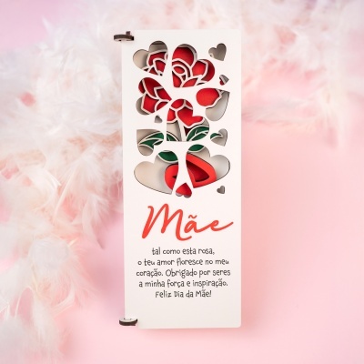 Cartão com recortes de corações e rosa vermelha, texto em português dedicado à mãe, fundo rosa e penas brancas