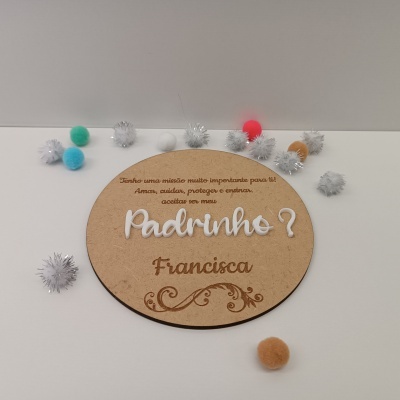 Placa redonda em madeira com texto Padrinho? e pompons coloridos ao redor.