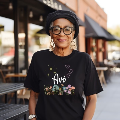 Pessoa com t-shirt preta com flores coloridas e texto Avó, chapéu e óculos, em ambiente urbano exterior.