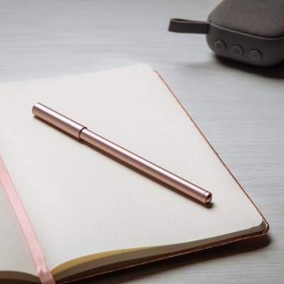 Caderno aberto com caneta metálica cor de rosa e coluna de som cinza ao fundo sobre mesa cinza clara
