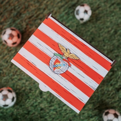 Caixa de cartão vermelha e branca com emblema de águia dourada, sobre relva artificial com bolas de futebol ao fundo