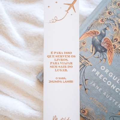 Marcador de livros branco com texto e desenho dourado posicionado sobre tecido branco felpudo ao lado de livro com capa azul e desenho de pavão.