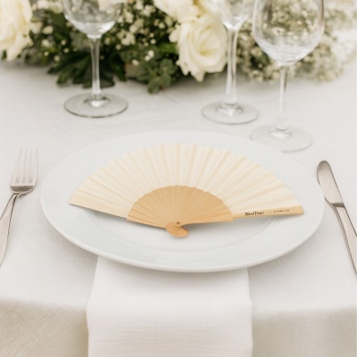 Leque de madeira e papel branco e bege sobre prato branco com decoração de mesa e flores