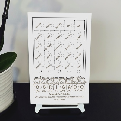 Poster branco com peças de puzzle e nomes, texto OBRIGADO e desenho de crianças, sobre suporte branco perto de planta