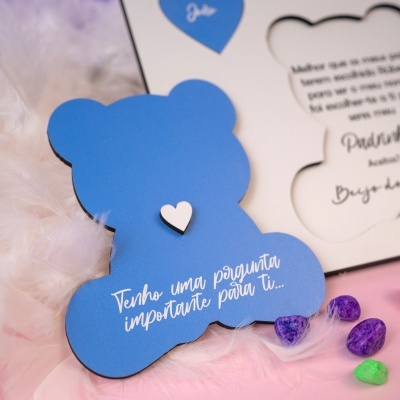 Cartão azul em forma de urso com texto em português sobre penas e pedras coloridas