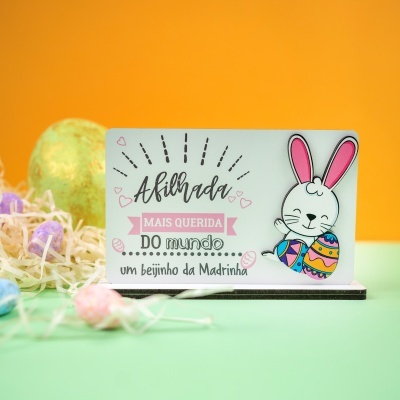 Placa decorativa com coelho e texto em português, sobre superfície verde com decoração de páscoa