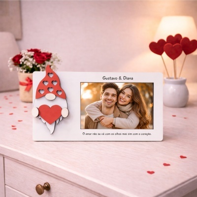 Porta-retrato branco com figura de gnomo de madeira segurando coração vermelho numa mesa clara com flores vermelhas e decoração de corações vermelhos