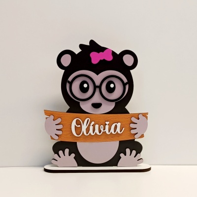 Figura de urso preto e cinzento com laço cor-de-rosa segurando placa de madeira com nome Olívia