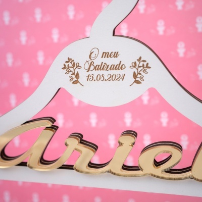 Placa em madeira branca e dourada com texto gravado O meu Batizado 15.08.2024 e fundo rosa com padrão.