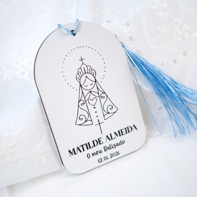 Etiqueta branca com desenho religioso, cordão azul e texto impresso sobre tecido branco bordado.