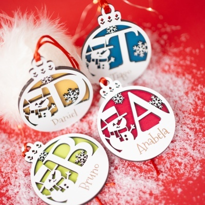 Ornamentos de Natal redondos coloridos com bonecos de neve e nomes, pendurados com cordões vermelhos em fundo vermelho com neve.