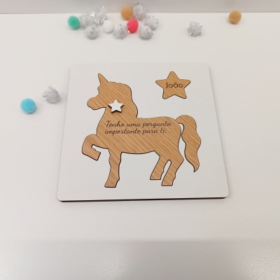 Cartão branco com recorte unicórnio e estrela em madeira com texto e nome, sobre mesa branca com pompons coloridos.
