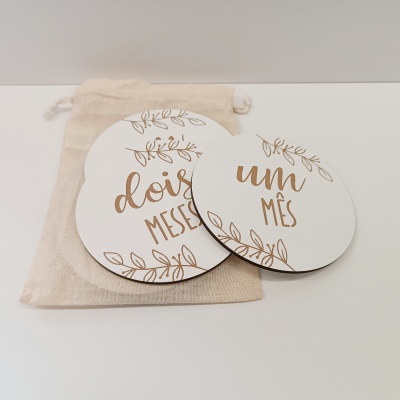Placas redondas em madeira branca com texto dourado e saco de tecido bege ao fundo
