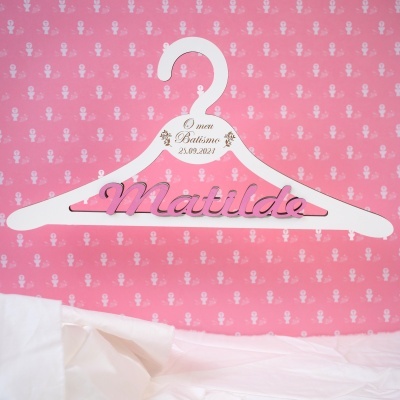 Cabide branco para bebés com texto Matilda e data de batismo, fundo rosa com padrão religioso.