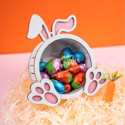 Embalagem de coelho branco com orelhas e patas cor-de-rosa cheia de ovos de chocolate coloridos sobre palha amarela e mini cenoura de plástico ao lado