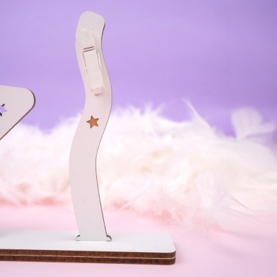 Suporte de madeira branco com prendedor e estrelas, sobre superfície branca e fundo lilás com plumas