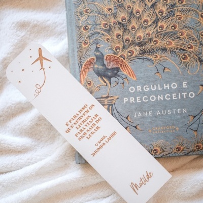 Livro com capa azul e pavão dourado e marca páginas branco com avião e texto em dourado