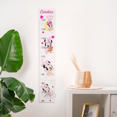 Régua de crescimento decorativa com desenho da Minnie pendurada numa parede branca ao lado de uma planta e uma mesa com vaso.