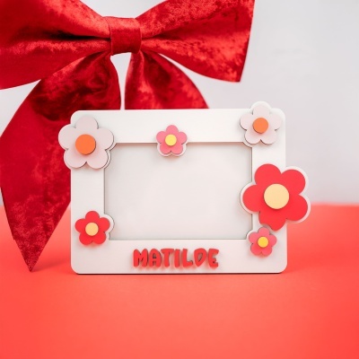 Moldura branca decorada com flores em tons de vermelho, rosa e cor-de-laranja, com o nome MATILDE em vermelho e laço vermelho ao fundo.