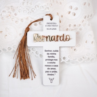 Cruz branca decorativa com nome Bernardo dourado e pendente castanho sobre tecido branco com rendas.