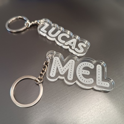 Porta-chaves transparente com nomes em branco LUCAS e MEL, argolas metálicas prateadas numa superfície preta