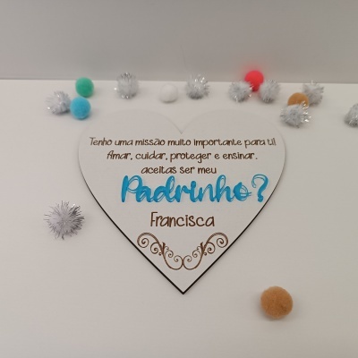 Placa de madeira branca em forma de coração com texto em castanho e azul, decorada com desenhos e pompons coloridos num fundo branco