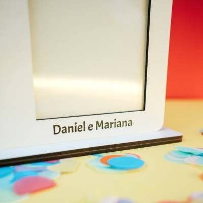 Objeto branco rectangular com moldura preta e texto Daniel e Mariana na base sobre superfície amarela com confetes coloridos