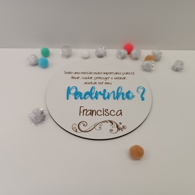 Placa redonda branca com texto em castanho e azul e pompons coloridos à volta sobre fundo branco
