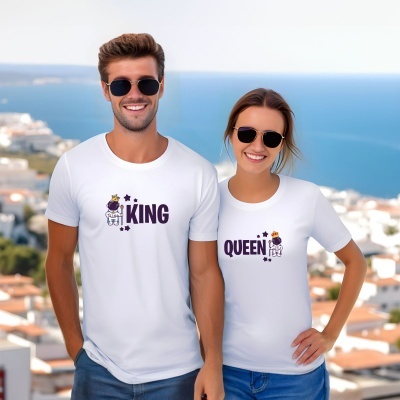Casal com t-shirts brancas KING e QUEEN com astronautas e coroas, ao ar livre com vista para o mar.