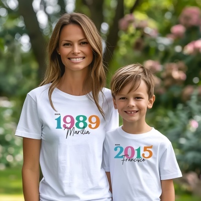 Duas pessoas vestem t-shirts brancas com números coloridos e nomes, em ambiente exterior com árvores e flores.