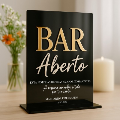 Placa preta de acrílico com texto dourado e branco colocada sobre mesa de madeira clara com flores ao fundo