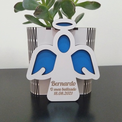 Figura de anjo branca com detalhes azuis e texto dourado em suporte preto, à frente de vaso branco com planta suculenta