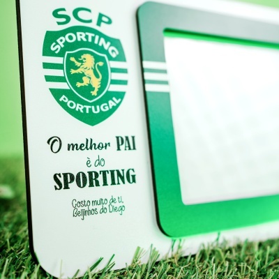 Moldura verde com placa branca com texto e logótipo do Sporting Portugal junto a relva artificial.