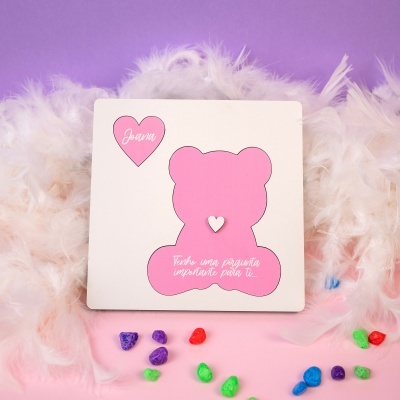 Cartão com urso cor-de-rosa e corações, penas brancas e pedras coloridas sobre fundo rosa e roxo.