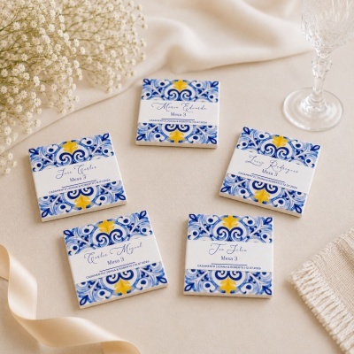 Peças de cerâmica decorativas com padrões azuis e amarelos e etiquetas de casamento numa mesa bege com flores e copo de cristal.