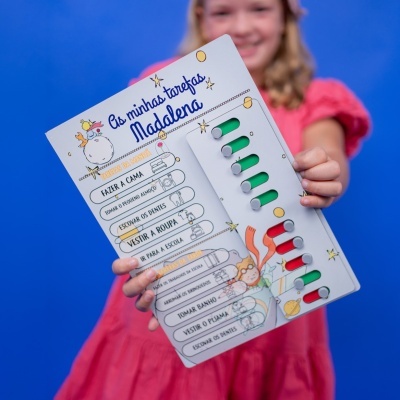 Quadro de tarefas infantil com listas e indicadores coloridos nas mãos de uma menina com vestido rosa