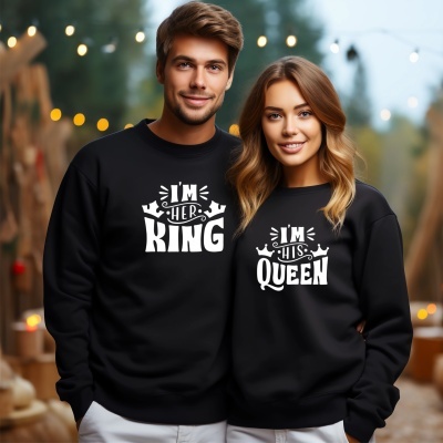 Casal com camisolas pretas com estampas IM HER KING e IM HIS QUEEN em ambiente exterior com luzes desfocadas.