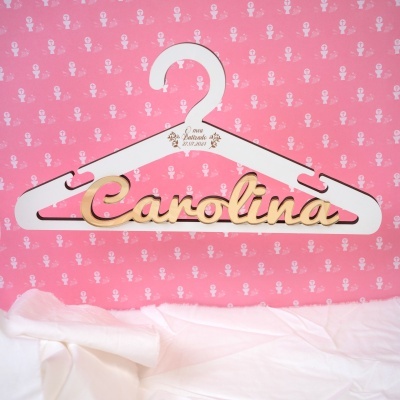 Cabide de madeira branco com nome Carolina dourado e inscrição O meu Batizado 27.07.2024, fundo rosa com símbolos religiosos brancos.