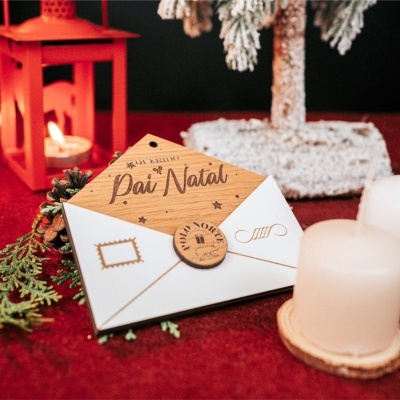 Cartão de Natal em madeira clara com texto Pai Natal, selo Polo Norte em envelope branco, castiçal vermelho com vela e decoração natalícia