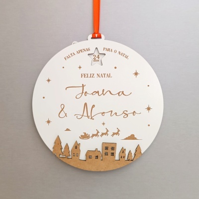 Ornamento de Natal redondo em madeira com texto e desenhos de vila e trenó voador.