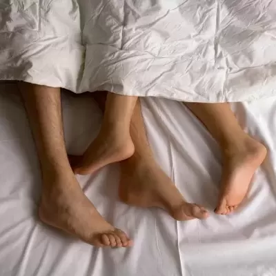 Pés de duas pessoas na cama com lençóis e edredom brancos.