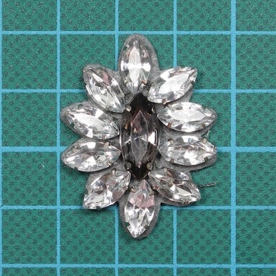 Broche com pedras cristal em padrão floral sobre fundo quadriculado verde.