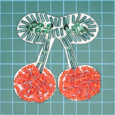 Aplicação em forma de cerejas com missangas verdes e vermelhas sobre fundo quadriculado azul