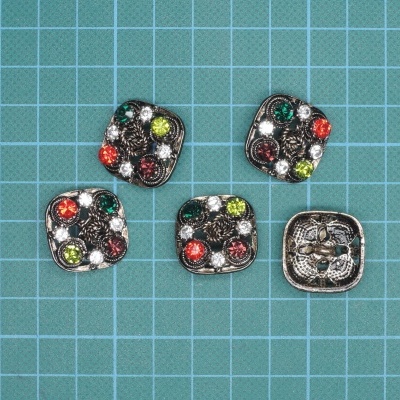 Cinco botões quadrados de metal prateado envelhecido com pedras coloridas sobre superfície de corte verde.